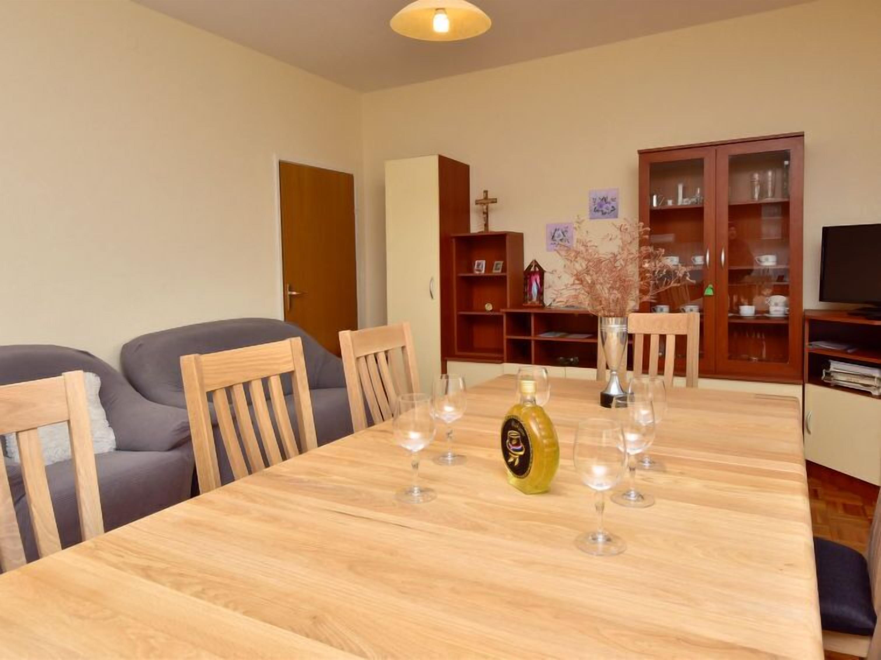 Un coin repas avec une table en bois, dressée pour six personnes, entourée de chaises en bois. On peut voir une bouteille d’huile d’olive et des verres à vin sur la table.