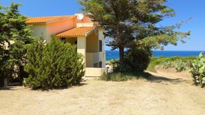 Un bâtiment à deux étages, doté d’un toit en tuiles, entouré de verdure et offrant une vue dégagée sur la mer.