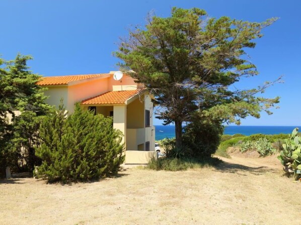 A two-story building with a tiled roof, surrounded by greenery and a clear view of the sea.