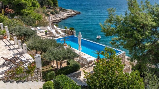 A pool area with a view of the sea, surrounded by greenery and stone structures.