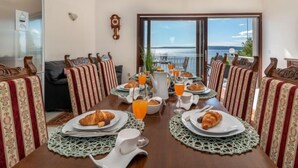 A dining area with a table set for breakfast, featuring croissants, orange juice, and a view of the sea.
