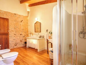 Un baño con pared de piedra, suelo de madera, lavamanos con espejo, inodoro y zona de ducha.