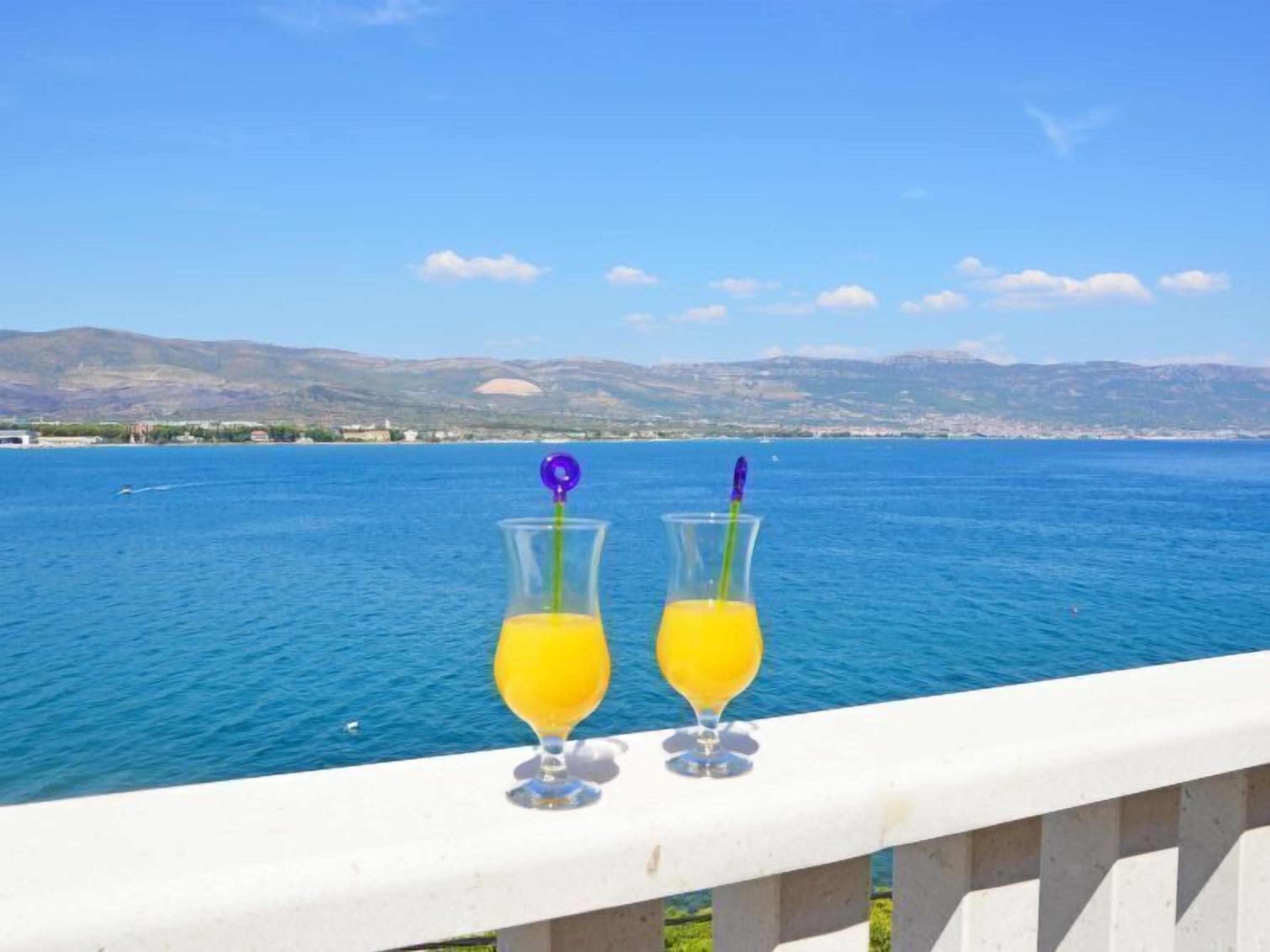 Two glasses of orange juice on a balcony with a view of a calm sea and distant mountains.