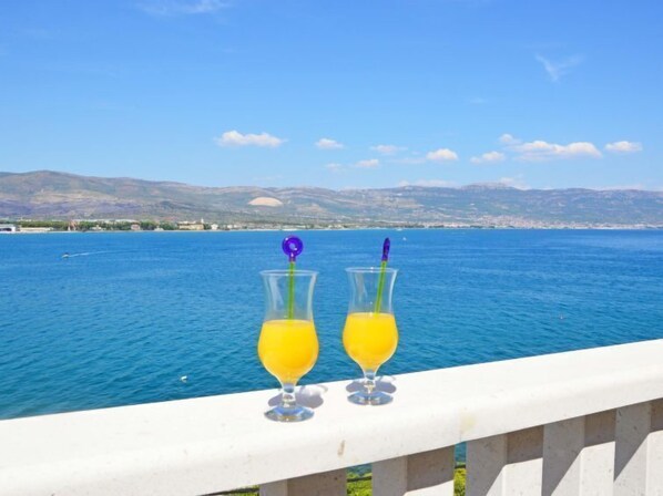 Dos vasos de jugo de naranja en un balcón con vista a un mar tranquilo y montañas lejanas.