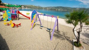 Une aire de jeux avec des balançoires et des toboggans, située près d’une plage où l’eau est d’un bleu limpide, avec des collines en arrière-plan.