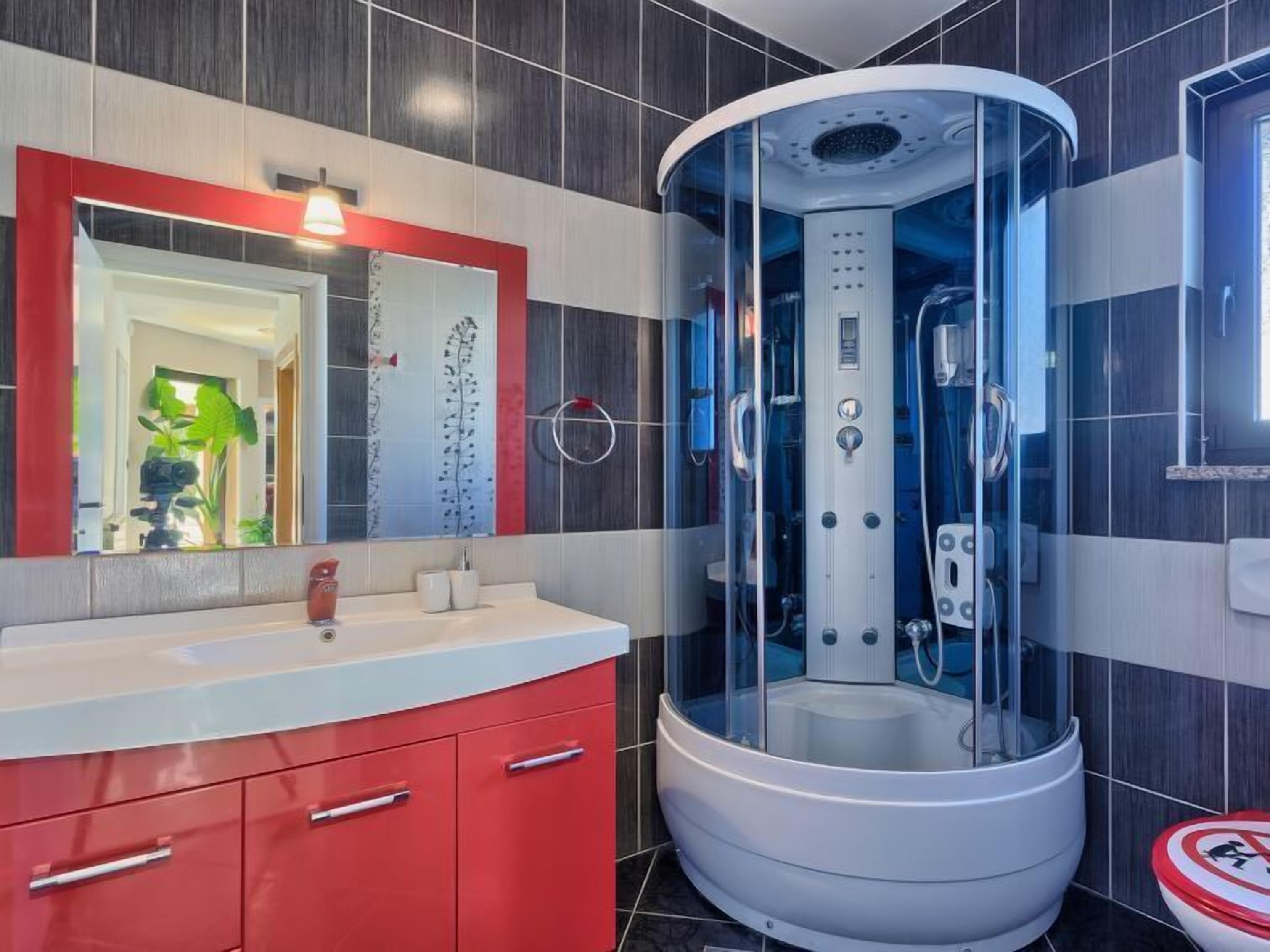 Un baño moderno con cabina de ducha de vidrio curvo, tocador rojo y un espejo grande con marco rojo.