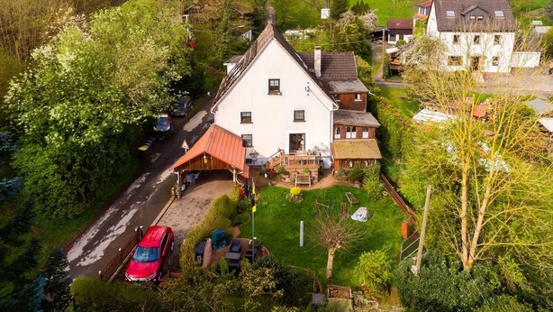 Ein weißes, zweistöckiges Haus mit rotem Dach, umgeben von üppiger Vegetation und einer kleinen Auffahrt.