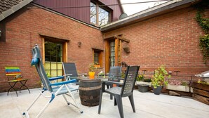 Eine Terrasse mit Backsteinmauer, einem Holztisch, zwei Stühlen, einem blauen Klappstuhl und Blumentöpfen.