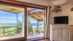 A room with a large sliding glass door leading to a balcony with a view of the sea. There is a wooden cabinet with a television on top, and an air conditioning unit mounted on the wall.
