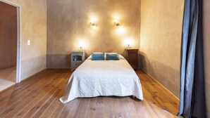 Une chambre avec un plancher en bois, un lit recouvert d’une courtepointe blanche, deux tables de chevet et des lampes fixées au mur.