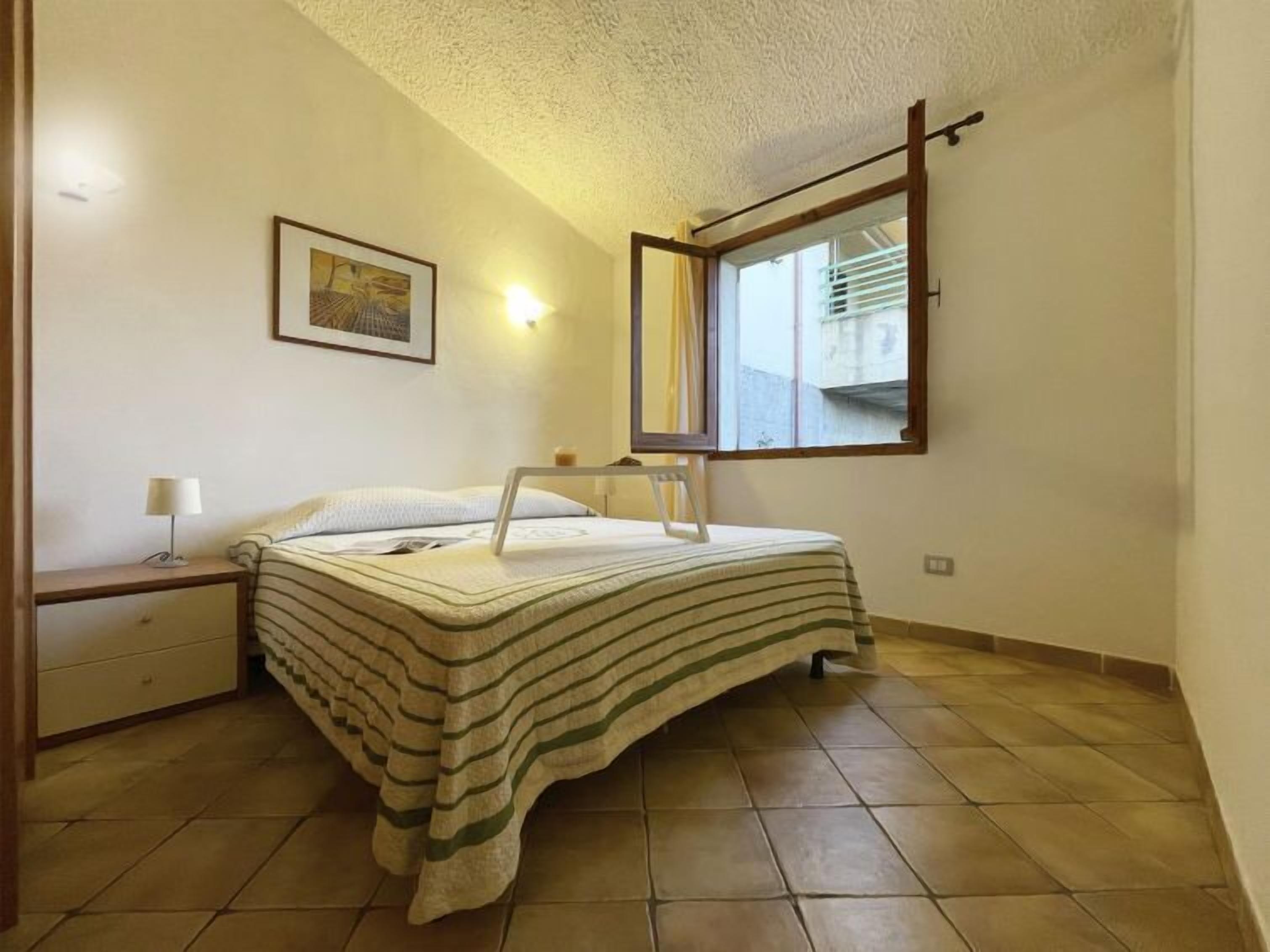 A bedroom with a bed, a nightstand, a lamp, a framed picture on the wall, and a window with a view of the outside.
