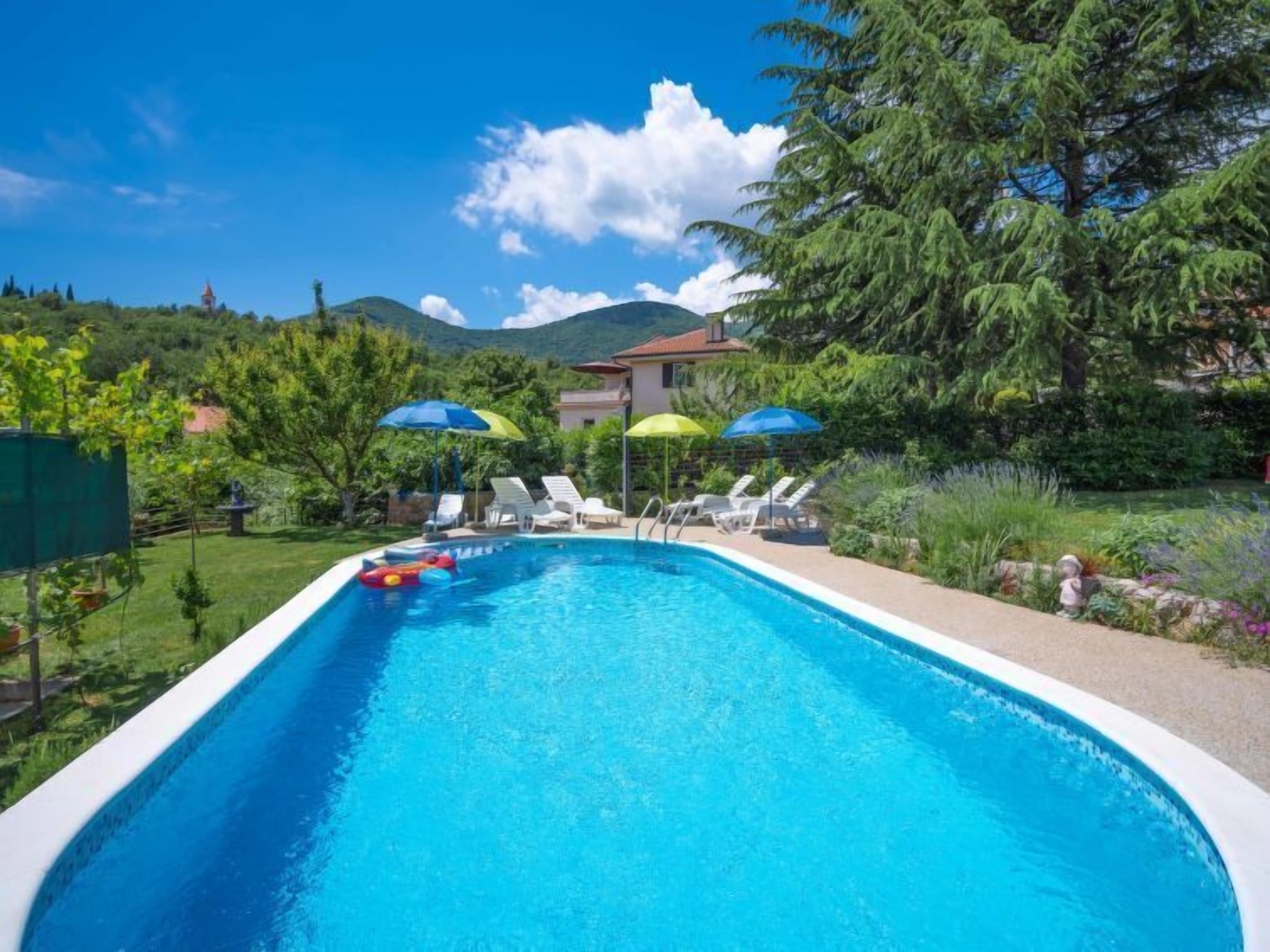 A swimming pool with lounge chairs and umbrellas, surrounded by greenery and mountains.