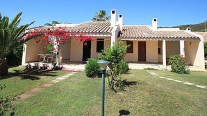 A two-story building with a tiled roof, a covered patio with outdoor furniture, and a garden with a palm tree and flowering plants.