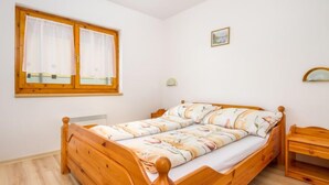 Un simple lit en bois, recouvert d’une couvre-lit à motifs floraux, se trouve dans une pièce aux murs blancs, avec une fenêtre ornée de rideaux et une petite table de chevet.