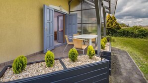 Une terrasse couverte avec une table et des chaises, une porte vitrée donnant sur l’intérieur et un jardin agrémenté de petits arbustes.