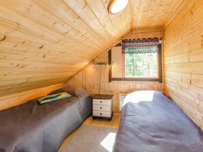 A small, cozy room with wooden walls and a sloped ceiling. There are two beds with matching bedding, a nightstand, and a window with a patterned curtain. - Lakunlahti by Interhome (Juuka)