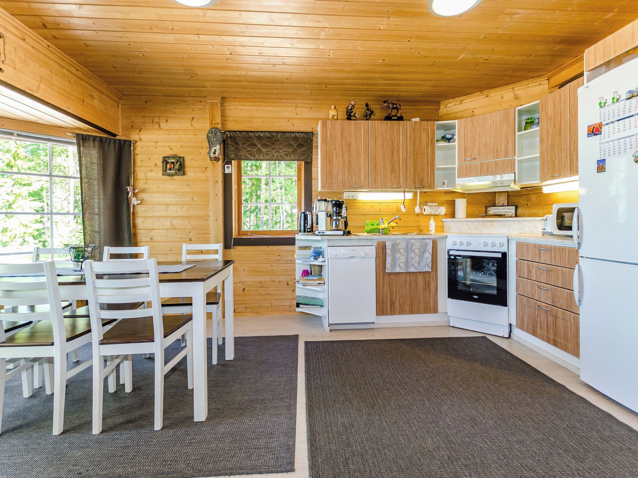 Interior d'una cabana de fusta amb taula de dinar, cadires, cuina amb fogó, forn i nevera, i una finestra amb vista a arbres.