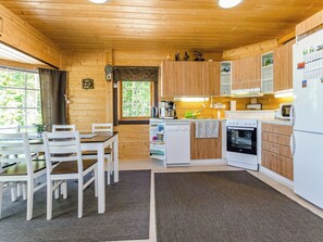 A wooden cabin interior with a dining table, chairs, a kitchen with a stove, oven, and refrigerator, and a window with a view of trees.