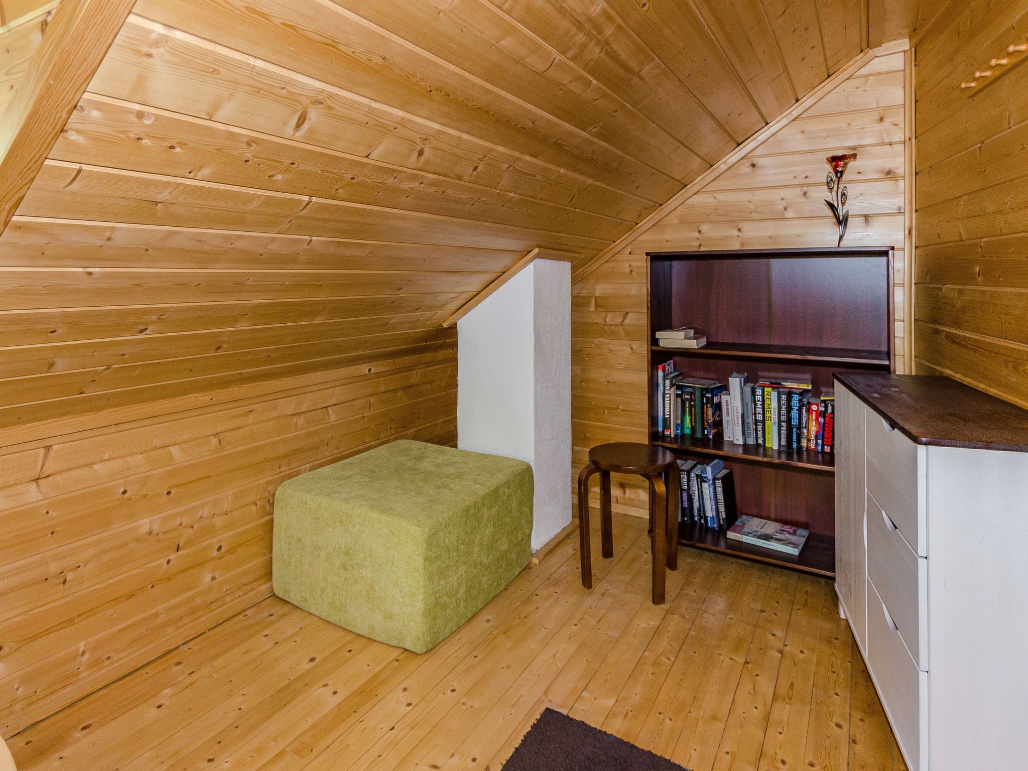 A cozy attic room with wooden walls and a sloped ceiling, featuring a green cushioned bench, a small wooden stool, and a bookshelf filled with books.