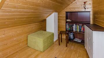 A cozy attic room with wooden walls and a sloped ceiling, featuring a green cushioned bench, a small wooden stool, and a bookshelf filled with books.