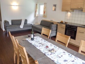 Un espace de vie moderne comprenant un coin repas, des canapés gris, des chaises en bois, une cuisine équipée d’une cuisinière et de placards, ainsi qu’une fenêtre avec des rideaux.
