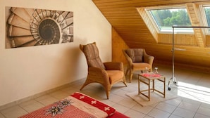 A cozy attic room with wooden beams, a large skylight, a painting of a spiral staircase, and two wicker chairs.
