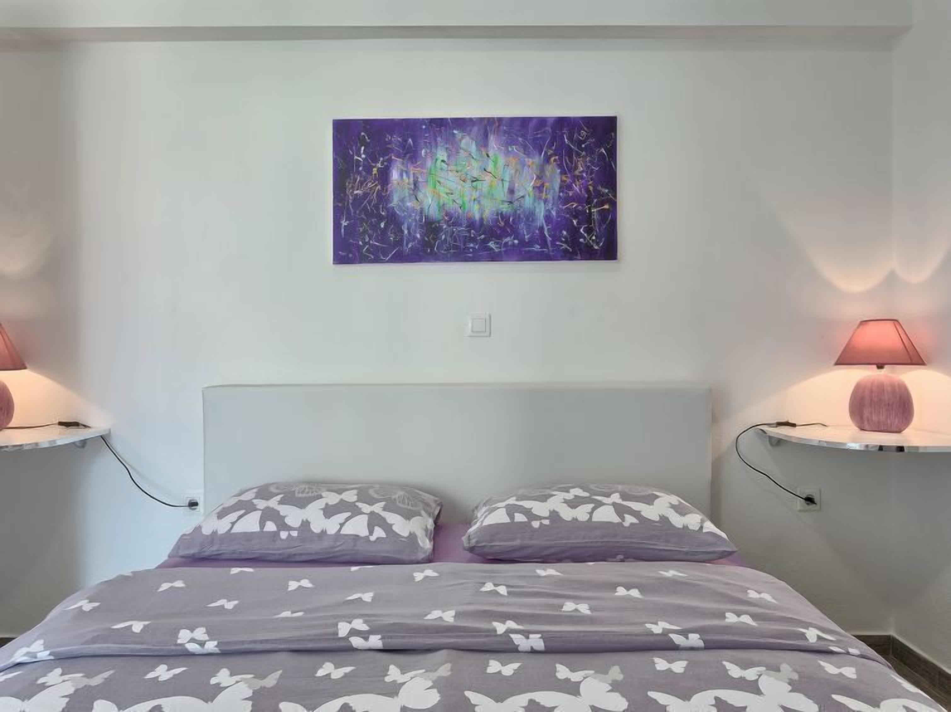 A bedroom with a bed, two bedside lamps, a wall-mounted abstract painting, and a white headboard.