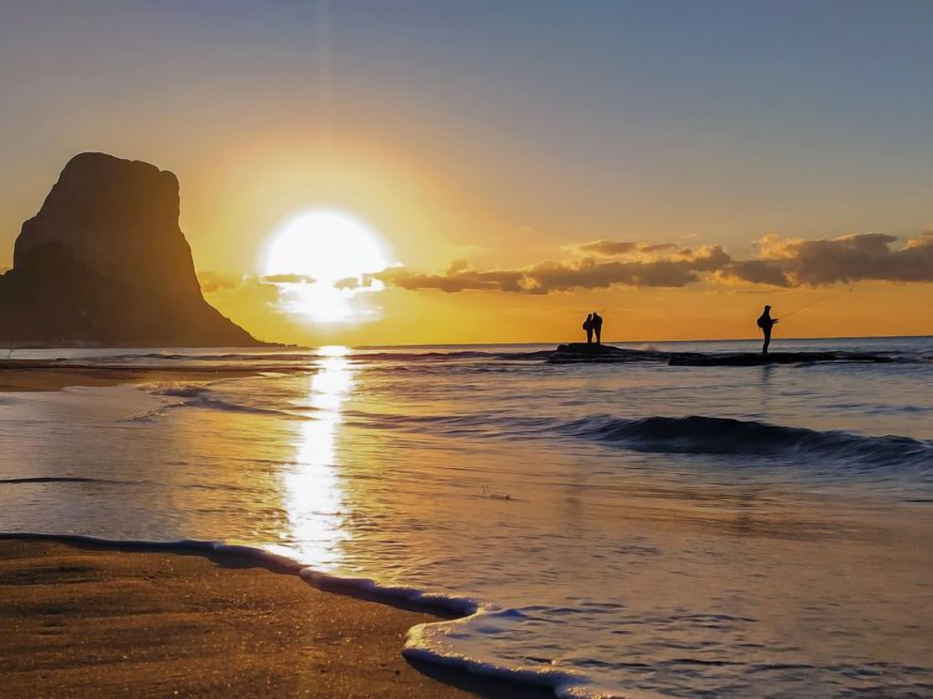 Eine Strandansicht mit einer großen Felsformation und untergehendem Sonnenuntergang über dem Meer.