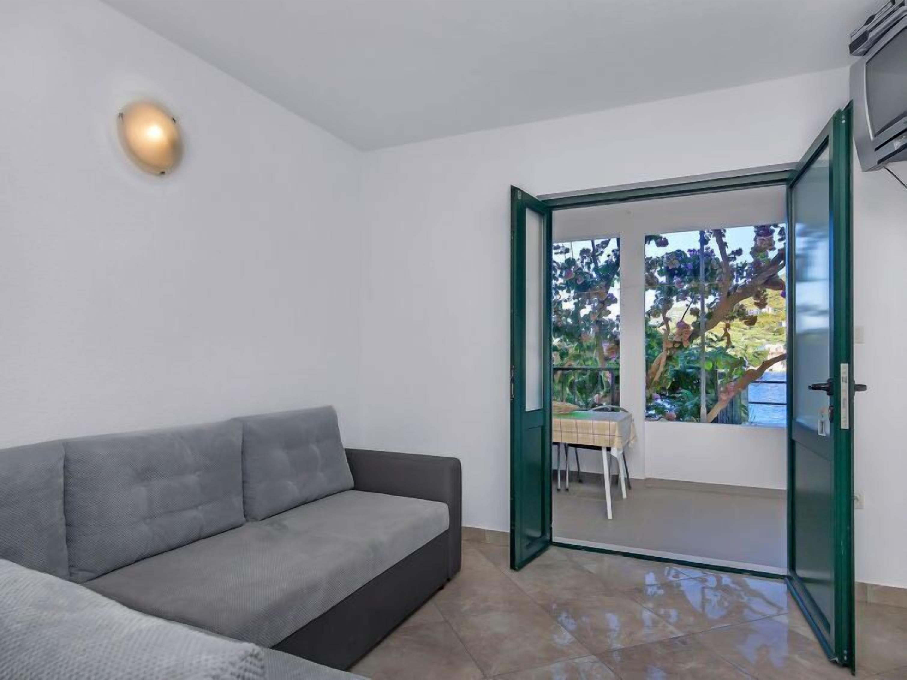 Un salon moderne avec un canapé gris, une porte vitrée donnant sur un balcon d’où l’on peut admirer la vue sur les arbres et une table.