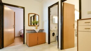 Une pièce avec une armoire en bois, un miroir, un petit meuble orné d’une plante, et une machine à laver en arrière-plan.