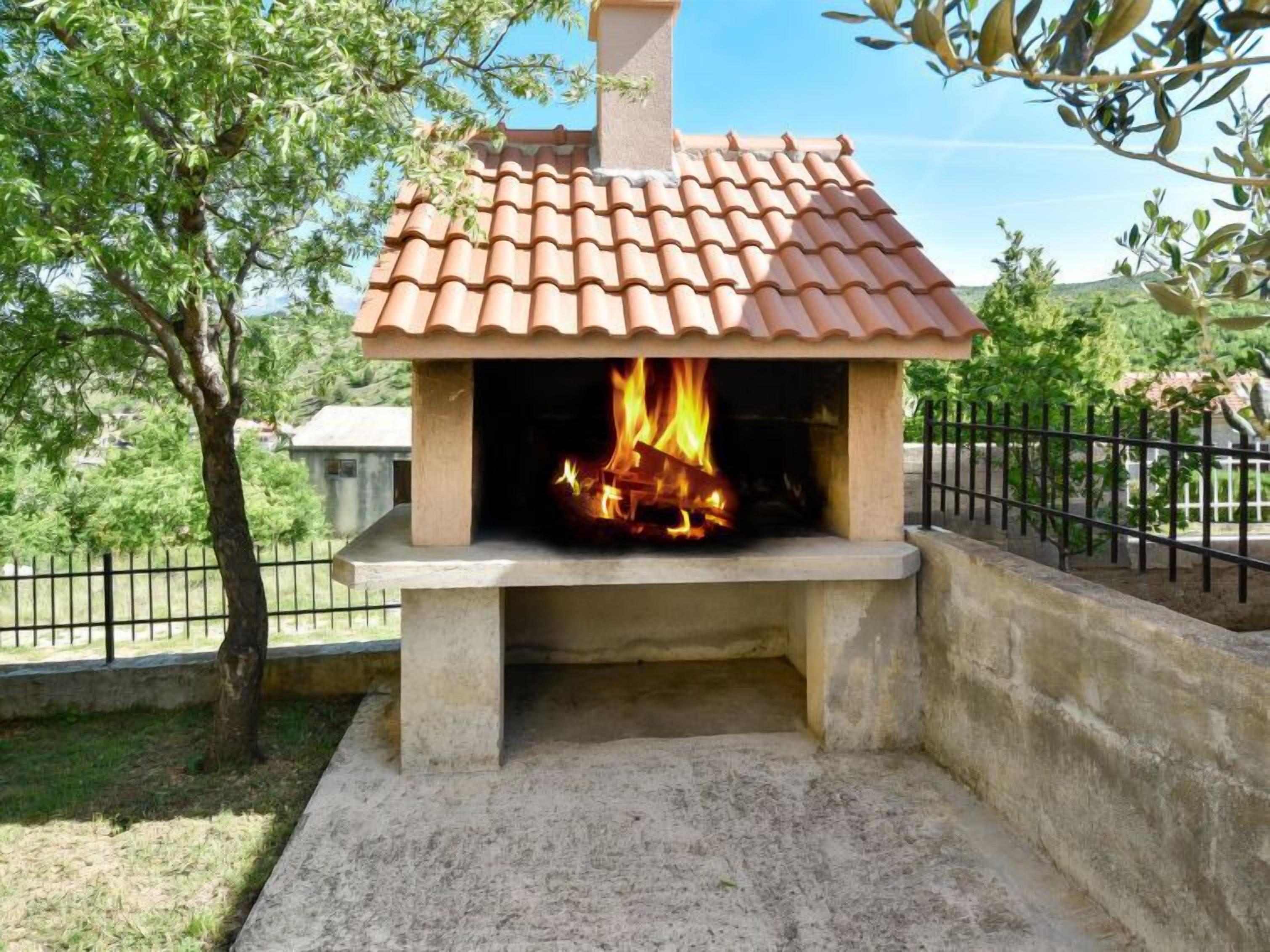 A stone-built outdoor pizza oven with a tiled roof, surrounded by a concrete base and a metal fence.