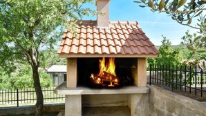 Un four à pizza extérieur en pierre, surmonté d’un toit en tuiles, entouré d’une base en béton et d’une clôture en métal.