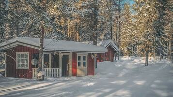 A small, single-story cabin with a porch, surrounded by snow-covered trees.