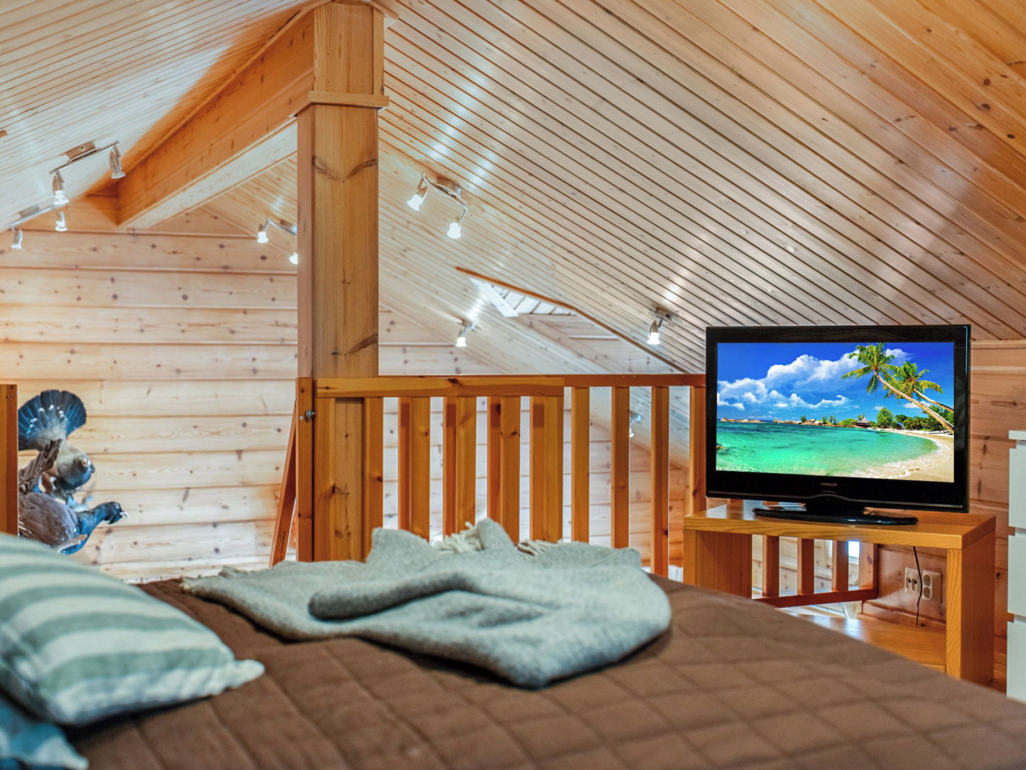 Una camera accogliente con soffitto in legno, un letto con coperta, una televisione su un mobiletto in legno e una ringhiera in legno.