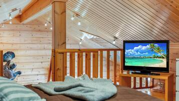 Una camera accogliente con soffitto in legno, un letto con coperta, una televisione su un mobiletto in legno e una ringhiera in legno.