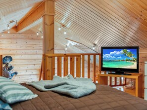Um quarto aconchegante com teto de madeira, uma cama com cobertor, uma televisão sobre suporte de madeira e um corrimão de madeira.