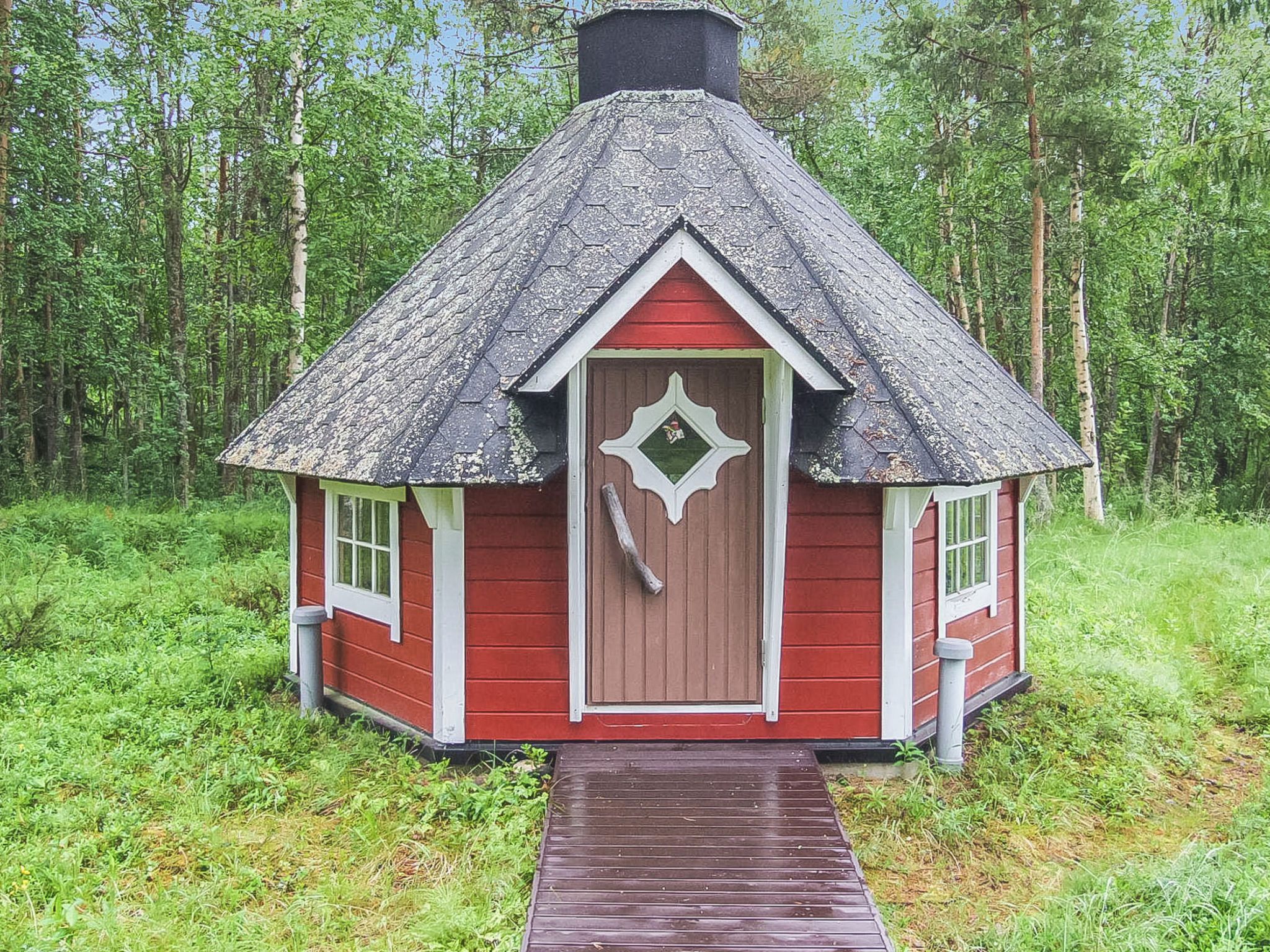 A small red cabin with a steeply pitched roof, a front door with a decorative window, and a wooden walkway leading to it.