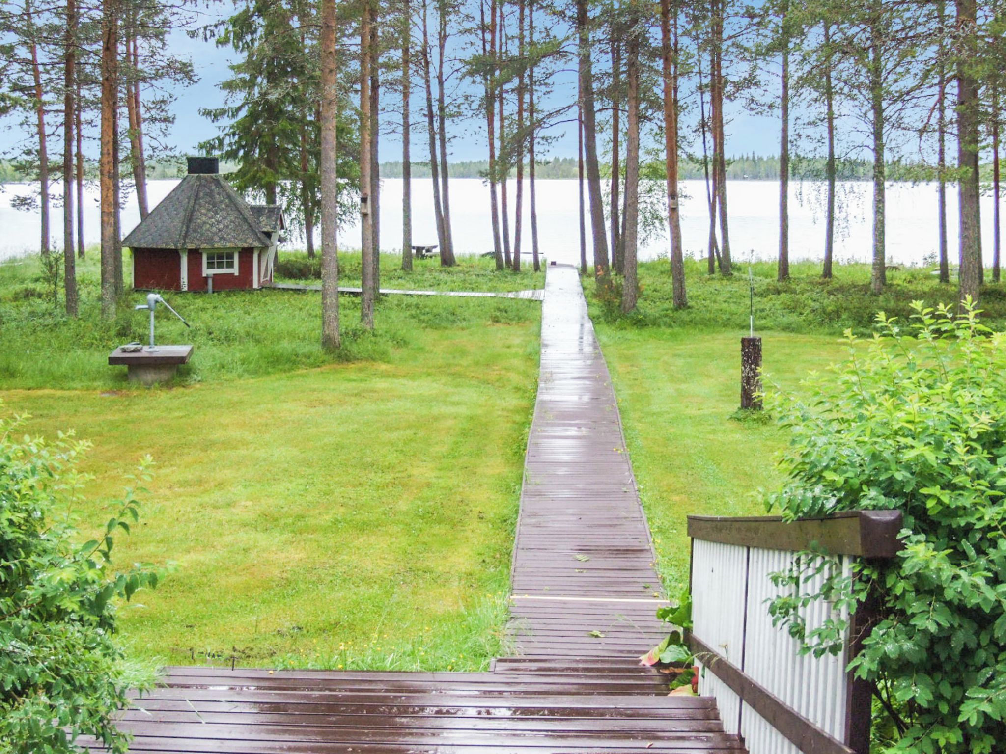 A wooden deck leading to a small house with a red roof, surrounded by tall trees and a body of water in the background.