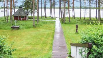 A wooden deck leading to a small house with a red roof, surrounded by tall trees and a body of water in the background.