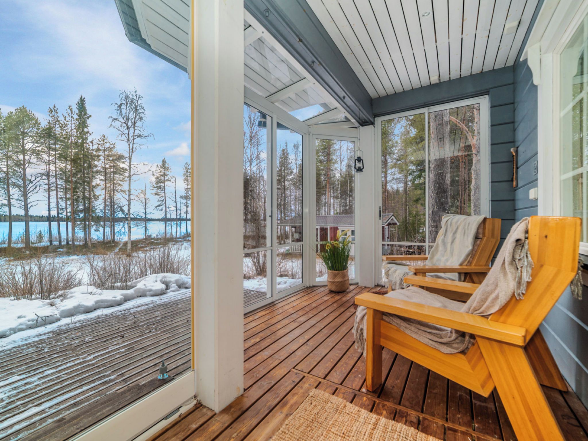 A covered deck with wooden flooring, two lounge chairs, a potted plant, and a view of a snowy landscape with trees and a body of water.