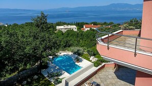 Una piscina con vista al mar y a las montañas, rodeada de vegetación y un edificio con balcón.