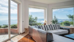 Una sala moderna con sofá y cama, ventanas amplias con vista al mar y a la ciudad, y piso de madera.