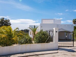 A modern building with a white facade, a gated entrance, and lush greenery around.
