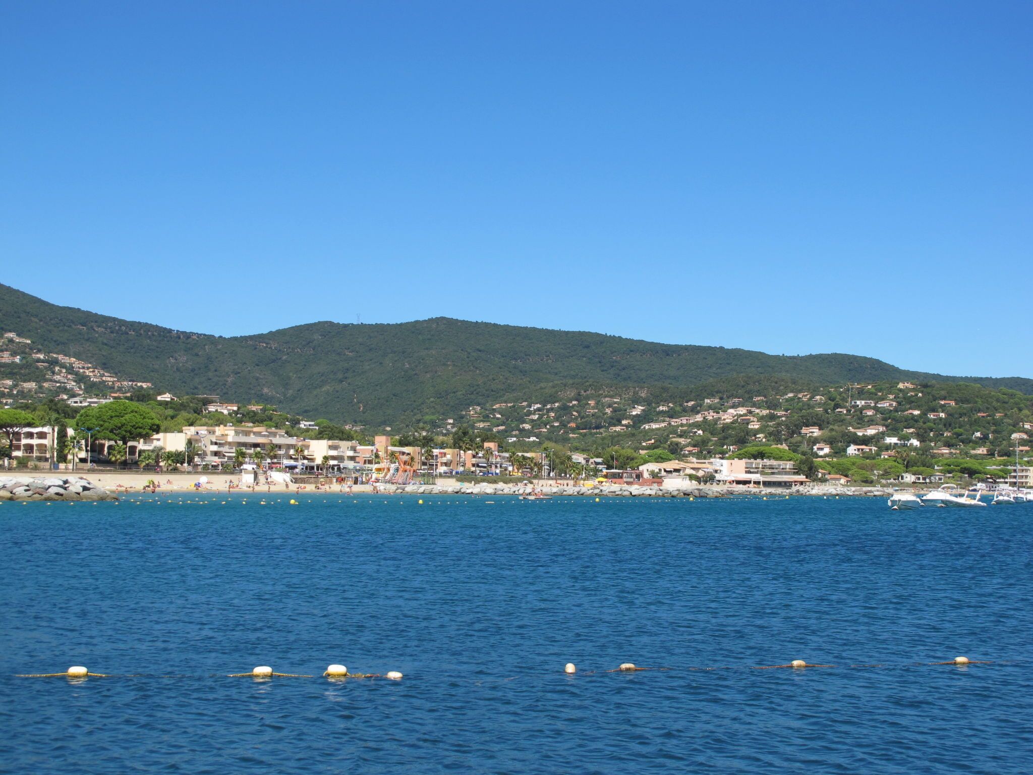 A coastal town with buildings along the shore, a clear blue sky, and a hilly backdrop.