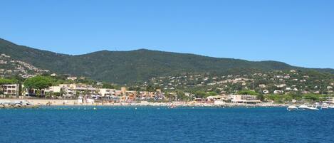 A coastal town with buildings along the shore, a clear blue sky, and a hilly backdrop.