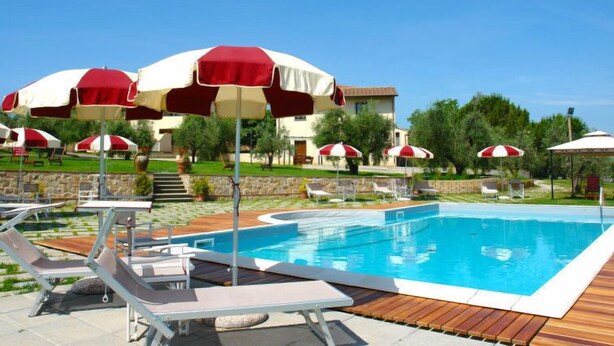 Une piscine entourée de chaises longues et d’ombrelles rouges et blanches, le tout bordé de verdure, avec un bâtiment visible au loin.