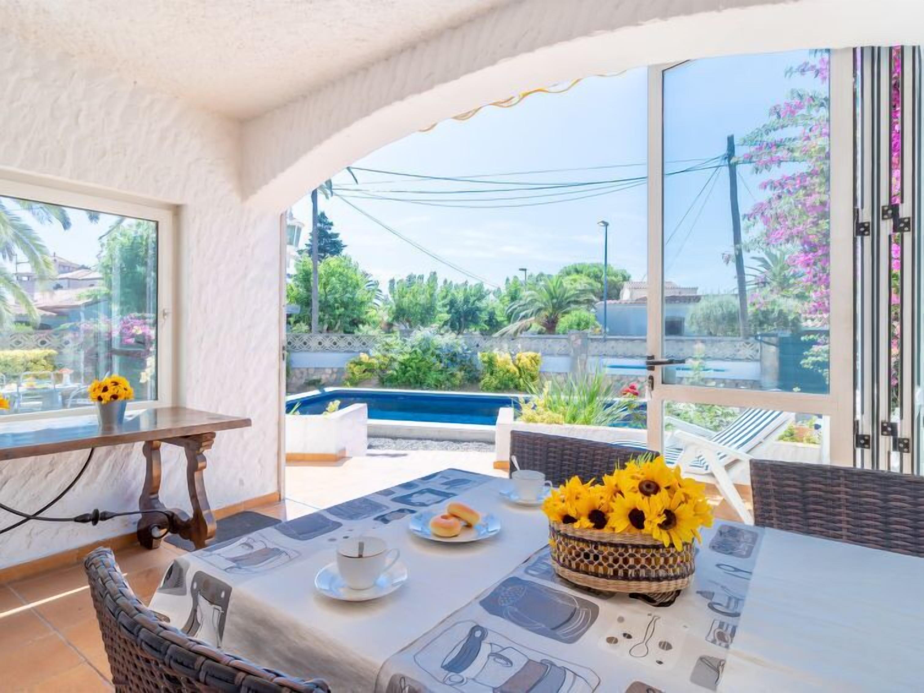 Zona pranzo con tavolo apparecchiato per due, un cesto di vimini con girasoli e vista su piscina e vegetazione all'esterno.