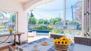 A dining area with a table set for two, a wicker basket with sunflowers, and a view of a swimming pool and greenery outside.