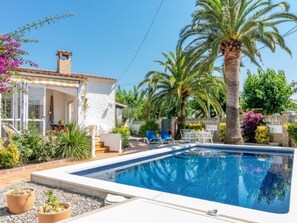 Un espace aménagé au bord d’une piscine à l’eau d’un bleu limpide, un bâtiment blanc à la façade en pierre et une végétation luxuriante comprenant des palmiers et des plantes à fleurs.