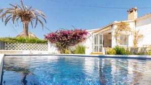 A swimming pool with clear blue water, surrounded by a white building with a glass door, a palm tree, and purple flowering plants.
